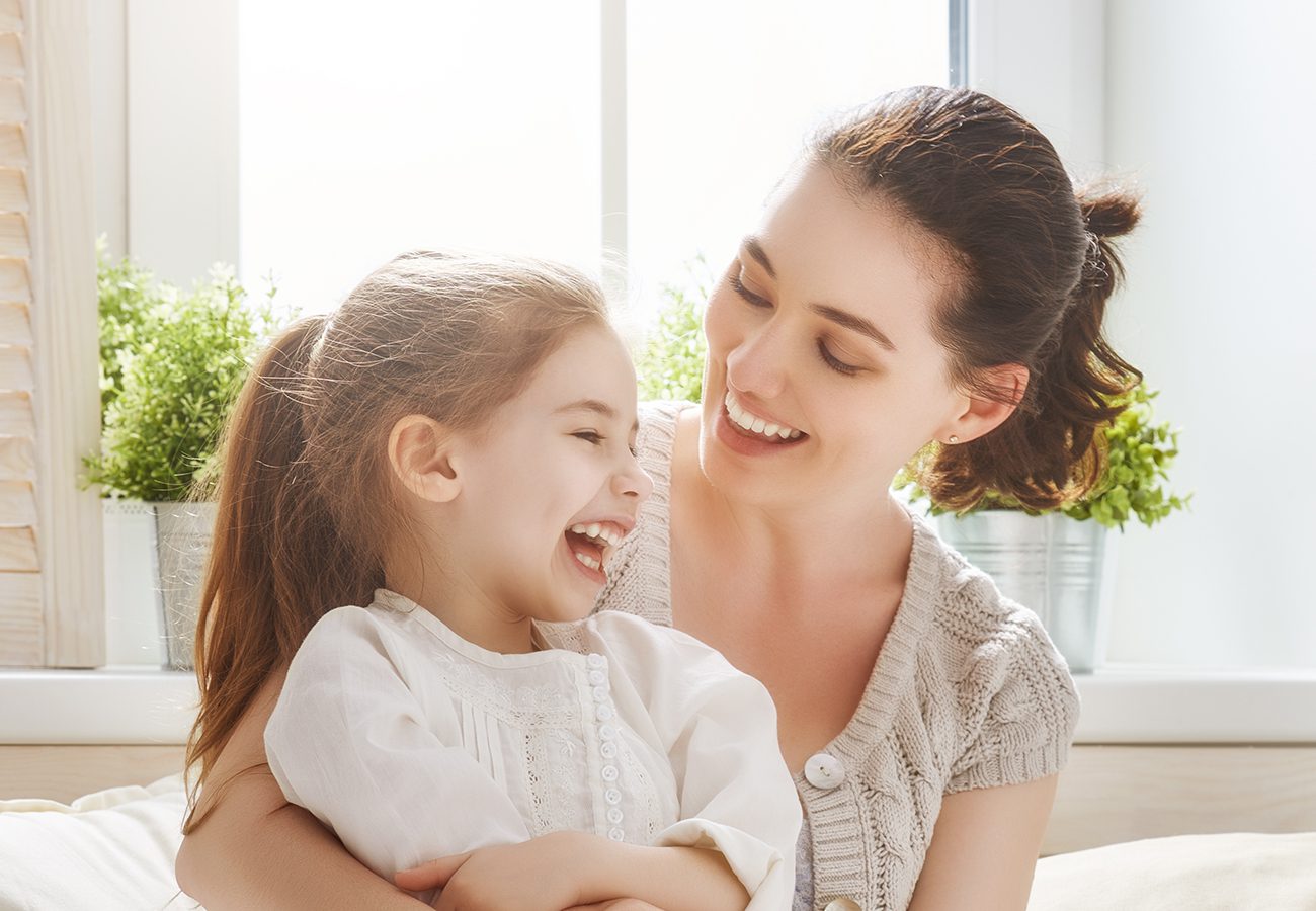 A woman and a young girl sit together indoors, smiling and laughing. Sunlight streams through a window behind them, with green potted plants on the windowsill. Both appear happy and relaxed.