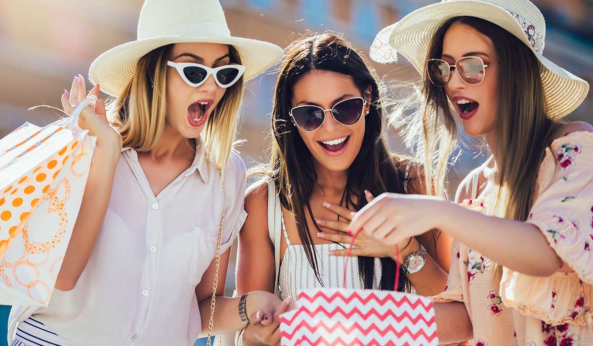 Three young women wearing sunglasses and sun hats excitedly look into shopping bags outdoors on a sunny day, smiling and appearing pleasantly surprised by their purchases.